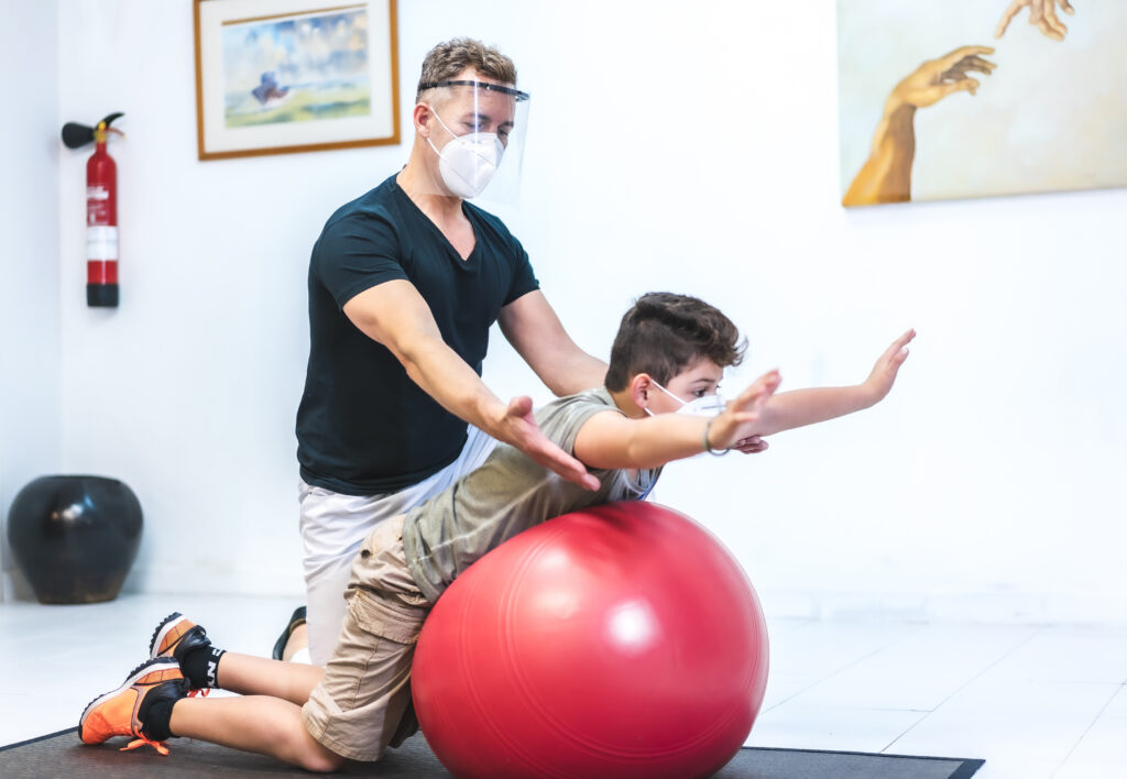 Physiotherapist with mask and screen stretching a child on a ball. Safety measures of physiotherapists in the Covid-19 pandemic. Osteopathy, therapeutic chiromassage