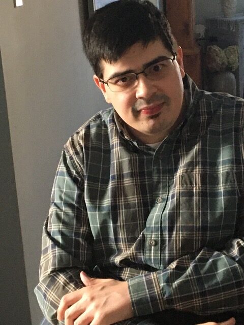 Scott Bremner, sitting in a chair posing for a photo wearing plaid shirt.