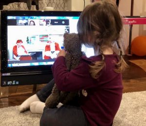 Little girl holding her stuffed animal close to her, sits in front of her computer on Zoom with Santa and Mrs. Claus.