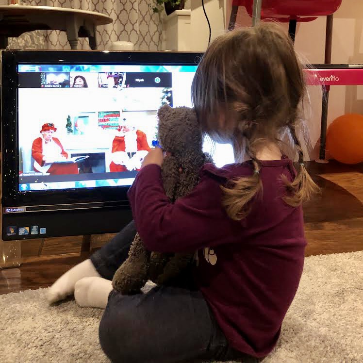 Little girl holding her stuffed animal close to her, sits in front of her computer on Zoom with Santa and Mrs. Claus.