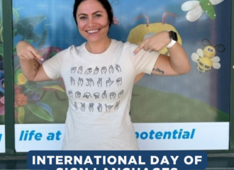 A female with dark hair, pointing to her white t-shirt which has sign language gestures.