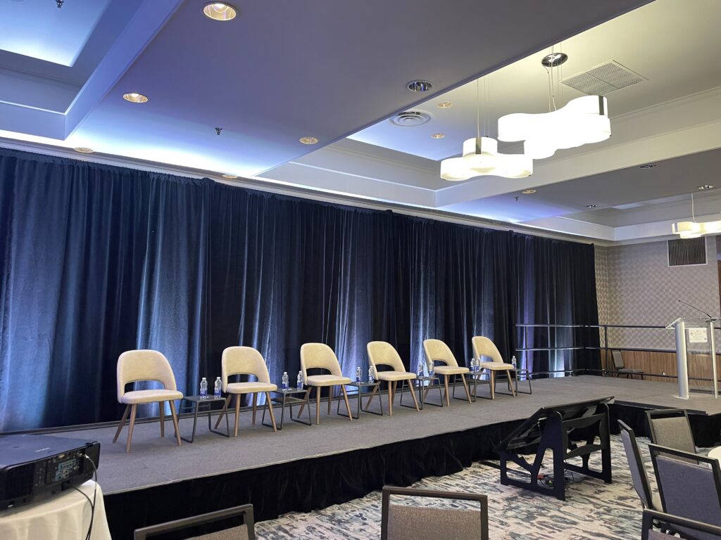 Photo of a stage with dark blue curtains and 6 white chairs facing the audience.