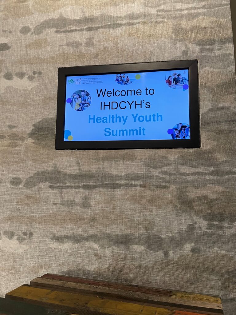 Photo of a grey wall with a television that has a screen which says "Welcome to IHDCYH's Healthy Youth Summit.