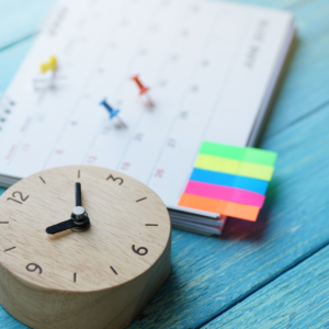 A small, wooden clock sits beside a calendar marked with colourful post-it notes and thumb tacks